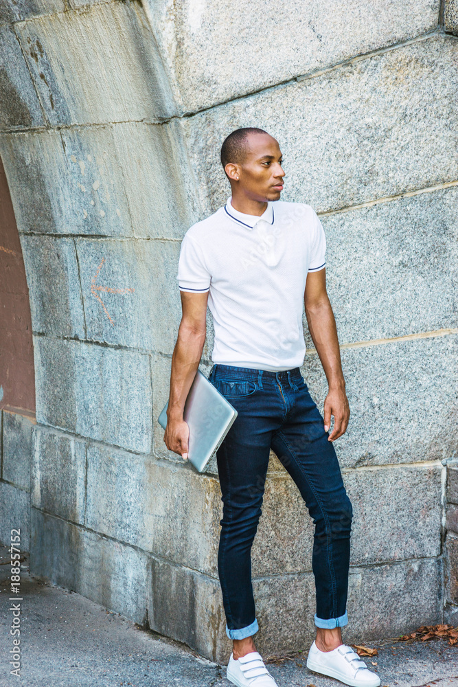African College Student studying in New York, wearing Polo shirt, blue jeans, white sneakers, carrying laptop computer, by rocky wall on campus, forward.. Photo | Adobe Stock
