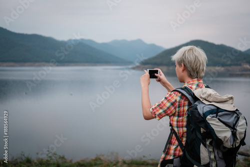 traveler hiker man with backpack hiking near lake. tourist backpacker use smart phone to take photo in the evening. travel lifestyle, summer vacation concept