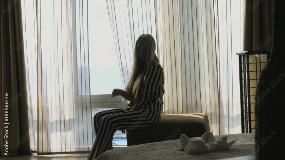 Young girl in a sexy striped suit departs from a panoramic window and sits down on the ottoman Young girl in a sexy striped suit departs from a panoramic window and sits down on the ottoman