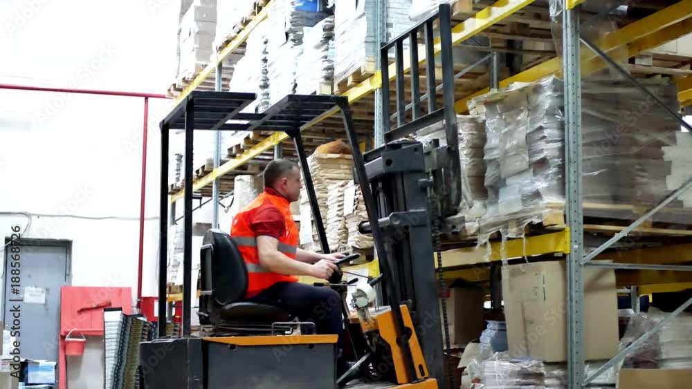 Warehouse worker driver in uniform moving cardboard boxes by forklift stacker loader. Forklift pallet with boxes in large modern warehouse Warehouse worker driver in uniform moving cardboard boxes by forklift stacker loader. Forklift pallet with boxes in large modern warehouse