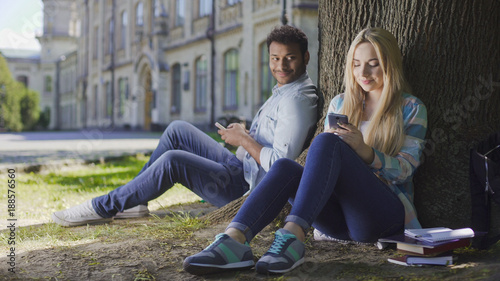 Man with cellphone sitting under tree and looking at girl using phone, affection