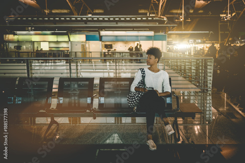 Beautiful young Black female tourist is sitting inside of modern airport terminal and waiting for her flight; cute Brazilian girl with curly Afro hair is sitting in waiting hall of railway station