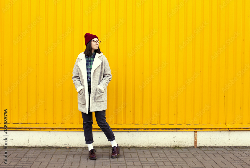 Millenial girl im fashionable clothes having a good time, making funny faces near bright yellow urban wall. Millenial girl im fashionable clothes having a good time, making funny faces near bright yellow urban wall.