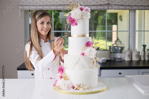 Beautiful smiling confident confectioner in chefs uniform standing in the modern kitchen, holding a brush and gold paint, decorating a four tier white, pink and gold wedding cake