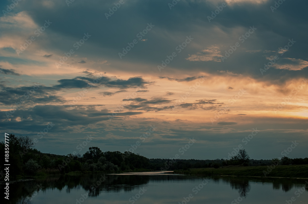 clouds and forest by the river on a meadow at sunset. Rural landscape.
