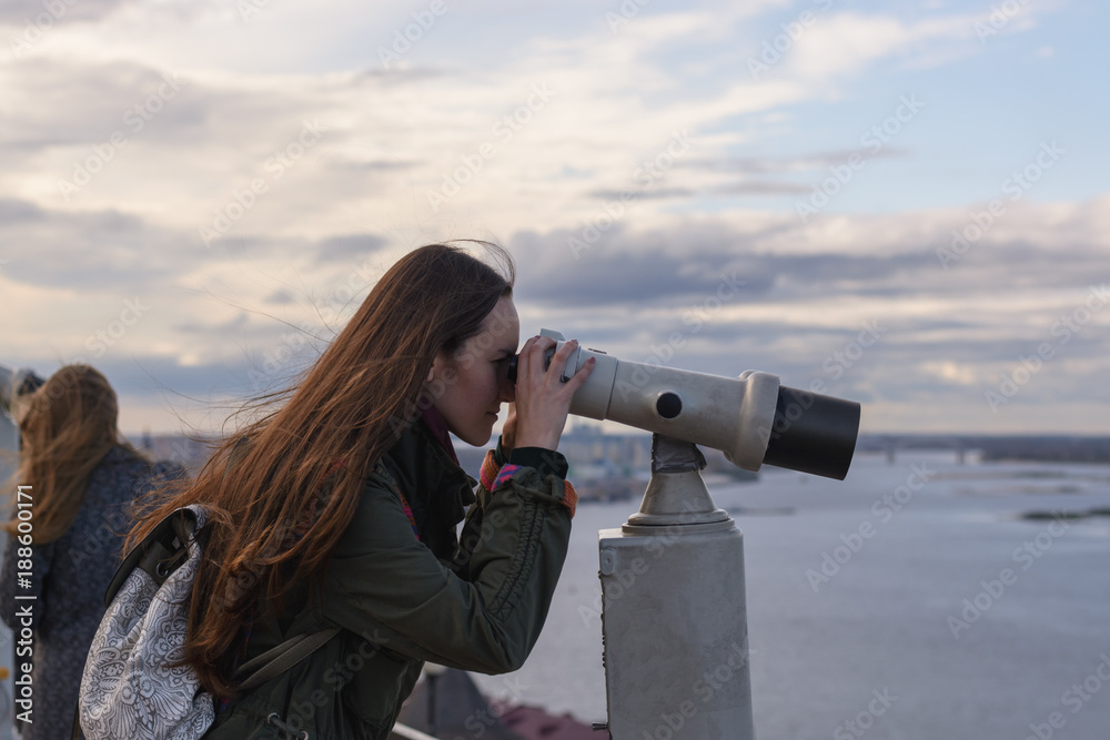 A young woman traveler on the observation deck looking through binoculars at the panorama of the city of Nizhny Novgorod