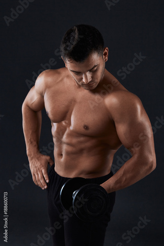 Muscular bodybuilder doing exercises with dumbbell over black background.Strong athletic man shows body,abdominal muscles,biceps and triceps.Work out,gaining weight,pumping up muscles with dumbbells.