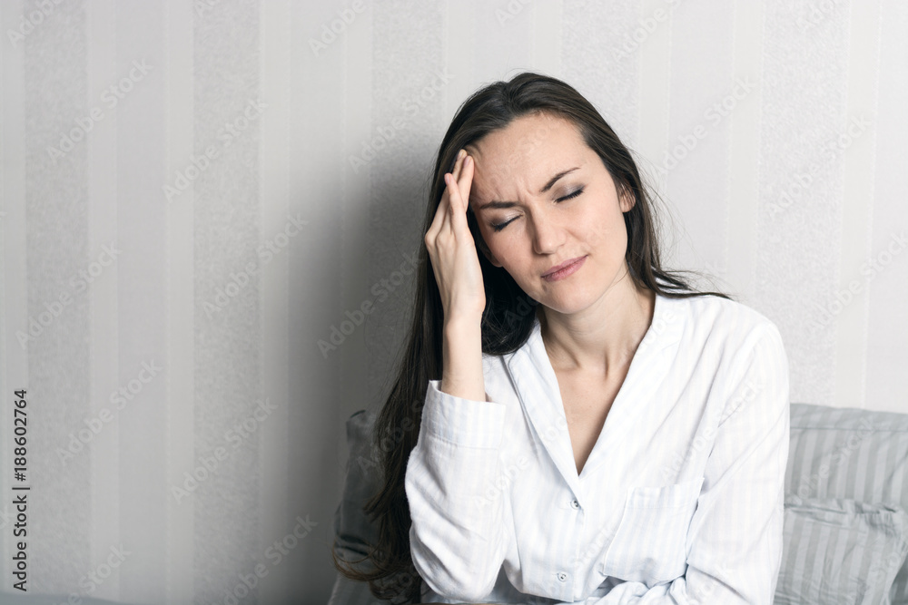 Portrait of a young brunette in pajamas sitting in bed, headache, migraine Portrait of a young brunette in pajamas sitting in bed, headache, migraine