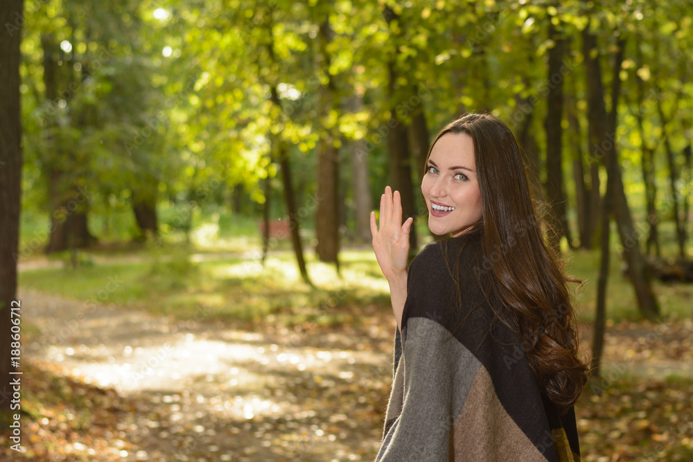Young brunette woman in a wool poncho standing in the sun on a footpath in a Park green and waving in greeting. Young brunette woman in a wool poncho standing in the sun on a footpath in a Park green and waving in greeting.