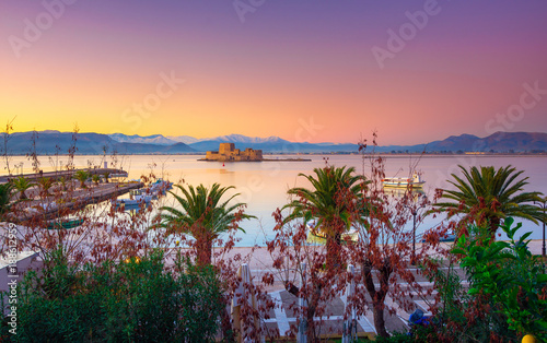 Beautiful port of Nafplio city in Greece with small boats, palm trees and Bourtzi castle on the water at sunset.
