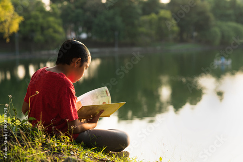 young asian boy in red shirt reading a book near lake garden in evening sun golden hour
