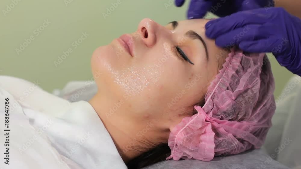 Cosmetologist applies cream on face of lying woman. Girl in bonnet lying on the session at the beautician. Woman receiving spa treatment Cosmetologist applies cream on face of lying woman. Girl in bonnet lying on the session at the beautician. Woman receiving spa treatment