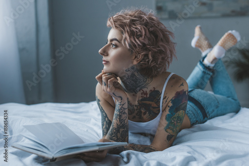 thoughtful tattooed girl lying on bed with book photo