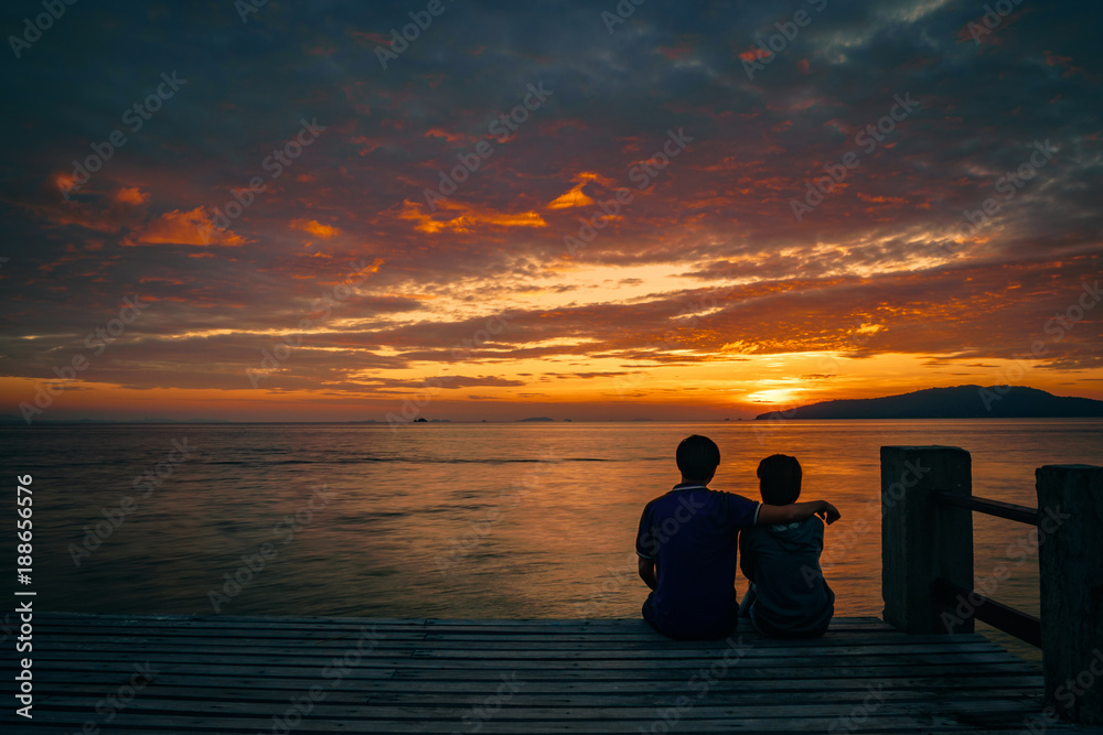 Silhouette of young romantic couple in love is sitting and hugging on wooden pier at the beach in sunrise time with golden sky. Vacation and travel concept. Romantic young couple dating at seaside.