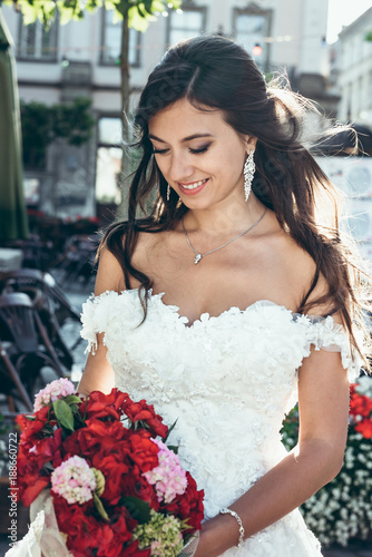 Outdoor portrait of the adorable brunetter bride with pretty smile is holding the wedding bouquet of red and pink flowers. photo