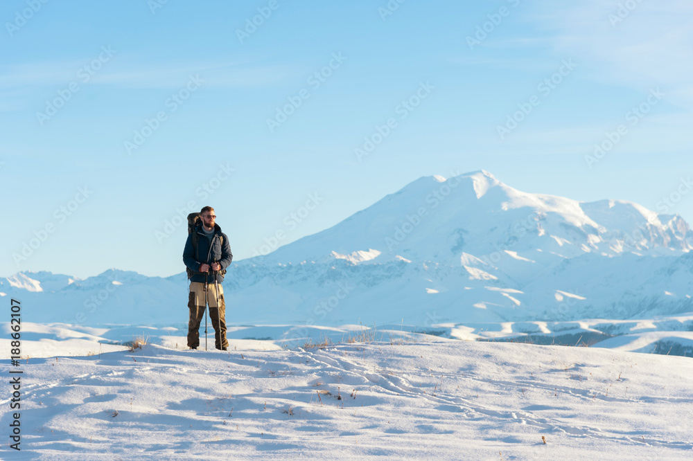 A traveler with a large backpack on his shoulders Stand on a snow-capped hill against the blue sky and the sleeping Elbrus volcano surrounded by the main Caucasian ridge and meditates.