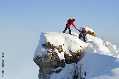 two hikers on top of the mountain in winter; a man helps a woman to climb a sheer stone photo