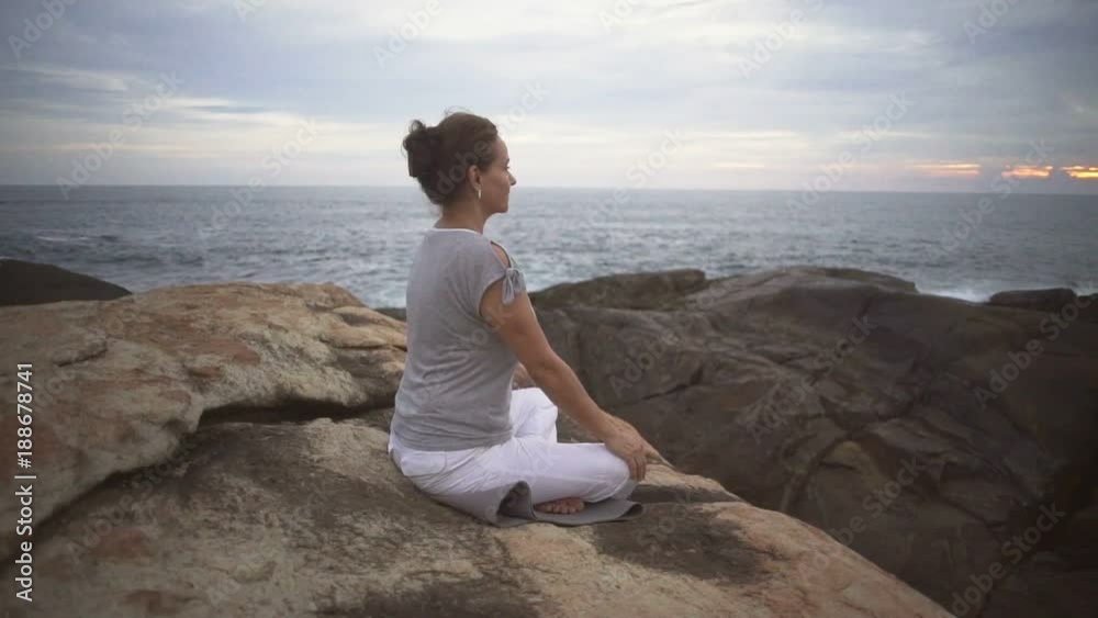 Woman meditate sitting on stone on rocky beach sunset rapid slow motion Woman meditate sitting on stone on rocky beach sunset rapid slow motion