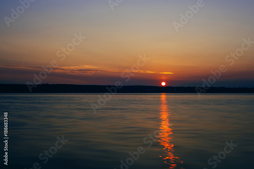 sunset over river with sun and silhouette of the shore on warm summer evening