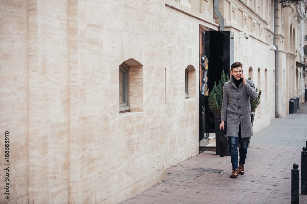 Photo of elegant male in coat with bag in hand walking down empty street, and talking on smartphone