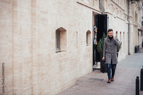 Photo of elegant male in coat with bag in hand walking down empty street, and talking on smartphone © Drobot Dean