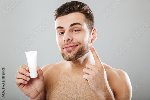 Close up image of handsome half-naked man with healthy skin applying face cream after shaving isolated over gray background