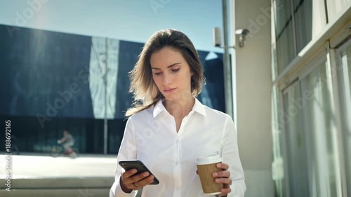 Beautiful young businesswoman wearing white shirt and using modern smart phone while walking at break in the city, professional female employer typing text message on cellphone outside, slow motion
