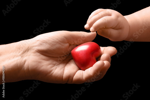 Baby taking small heart from hand of elderly man on dark background