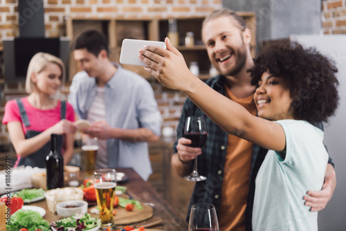 multiethnic friends taking selfie in kitchen with smartphone