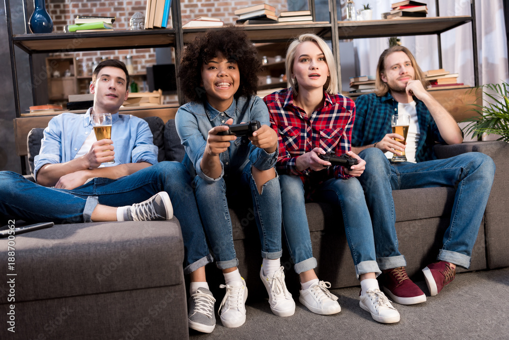 Obraz premium multicultural girls playing video game on sofa at home, men drinking beer