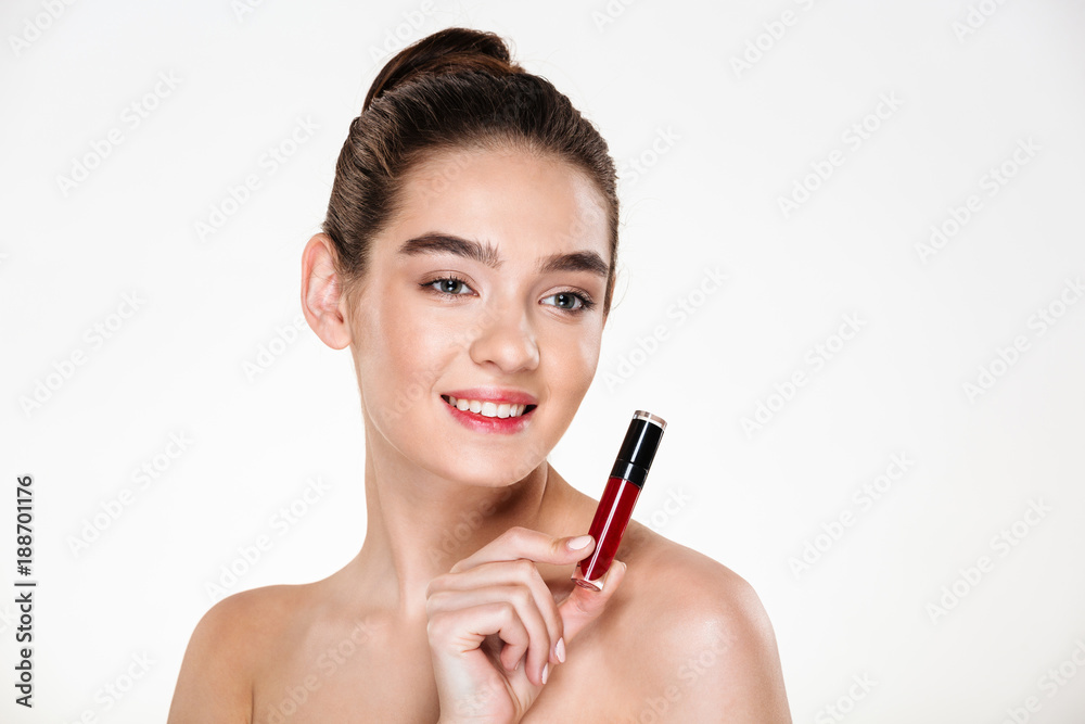 Close up studio portrait of smiling brunette woman wearing makeup holding lip gloss at her face and looking aside isolated over white background Close up studio portrait of smiling brunette woman wearing makeup holding lip gloss at her face and looking aside isolated over white background