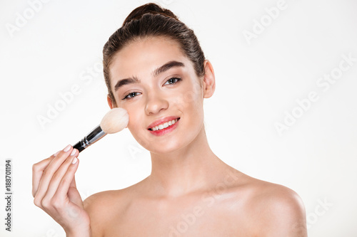 Beauty portrait of smiling half-naked woman with fresh skin applying makeup with soft brush and looking at camera isolated over white background