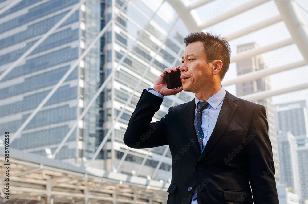 medium aged asian business man wearing modern black suit making phone call with mobile smart phone in building city background, internet network connection, technology communication, financial concept