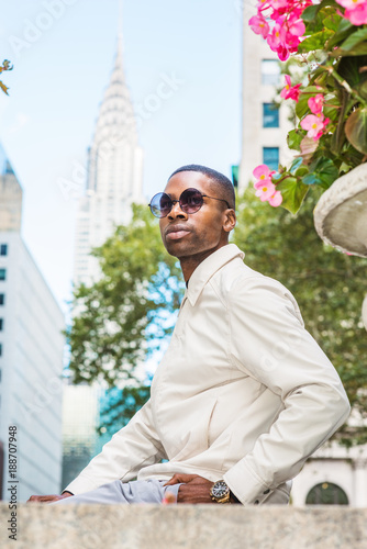 Young Handsome African American Man wearing sunglasses, looking around, relaxing outside in Manhattan, New York. Tall buildings, trees on background..