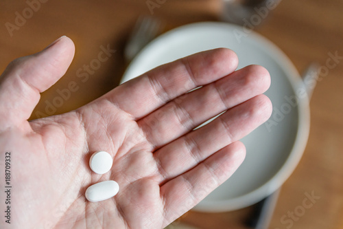 Close-up view of a white male hand holding two white pills in the palm above a blurry background showing a dining table with plate, cutlery and glass. photo