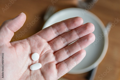 Close-up view of a white male hand holding two white pills in the palm above a blurry background showing a dining table with plate, cutlery and glass.