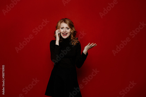 young stylish blond girl in black clothes posing in the studio isolated on red background, model tests, emotionally talking on a smartphone, laughing and smiling