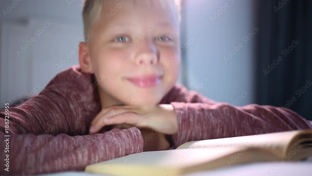 Closeup view of happy little 10 years old kid reading paper book with interest laying in his bed at home. Smiling cute child looks at camera happily. Closeup view of happy little 10 years old kid reading paper book with interest laying in his bed at home. Smiling cute child looks at camera happily.