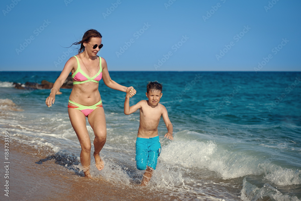 Mom and son are running along the sea shore. They are holding hands and moving towards the camera.