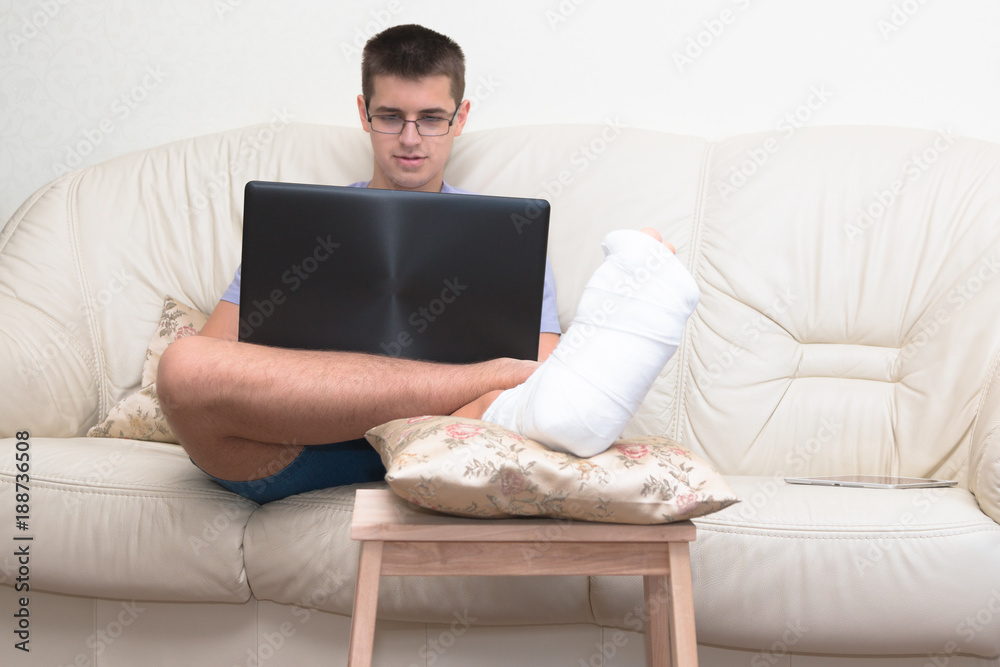 A teenager boy with a broken foot alone at home doing homework, reading. Leg in a cast. Disease. A teenager boy with a broken foot alone at home doing homework, reading. Leg in a cast. Disease.