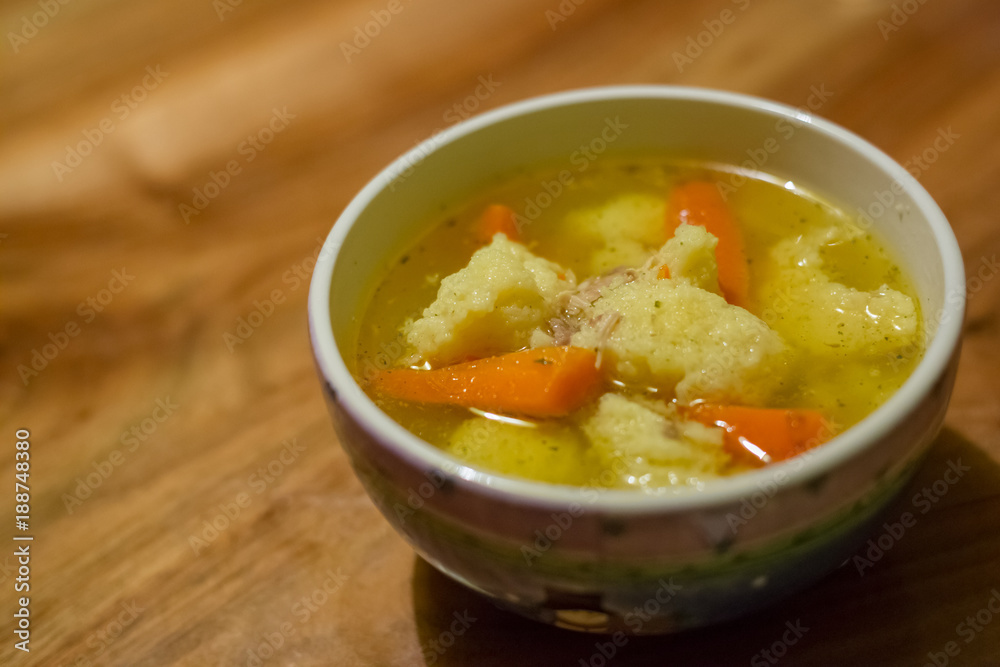 Hot chicken soup with dumplings and carrot in a small porcelain bowl on wooden table Hot chicken soup with dumplings and carrot in a small porcelain bowl on wooden table