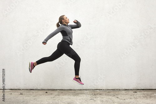 One young woman, jumping in mid-air, outdoors, white wall behind, simple minimalistic, sport clothes.