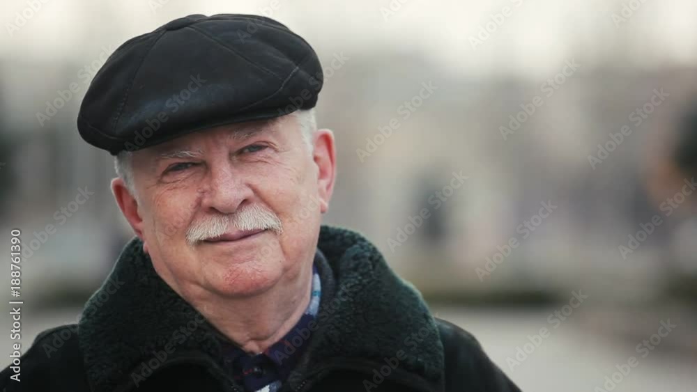 A cheery view of a senior man in a leather fur coat and a leather cap who stands on a city street and smiles full of Xmas spirit. A cheery view of a senior man in a leather fur coat and a leather cap who stands on a city street and smiles full of Xmas spirit.