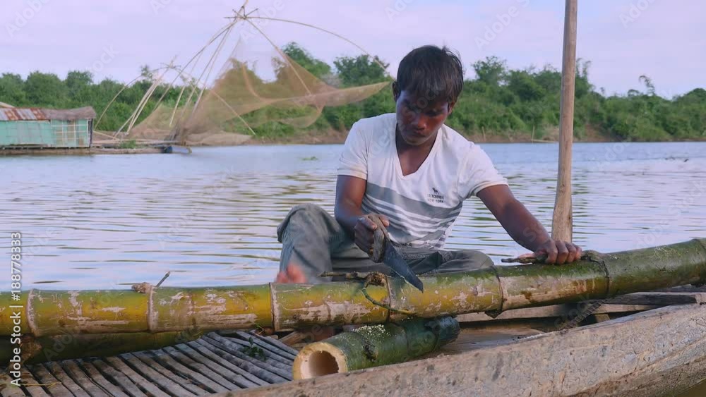 close up of a man sawing bamboo stalks in a dugout canoe on the riverside