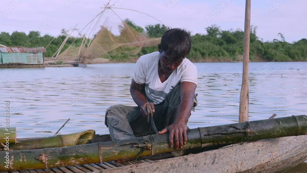 close up of a man sawing bamboo stalks in a dugout canoe on the riverside