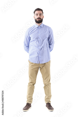 Front view of young smart casual bearded man standing with hands on back looking at camera. Full body length portrait isolated on white background. 