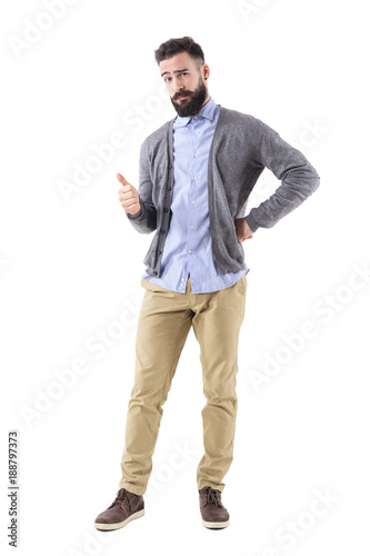 Stylish bearded smart casual guy wearing cardigan showing thumb up gesture and look at camera. Full body length portrait isolated on white background. 