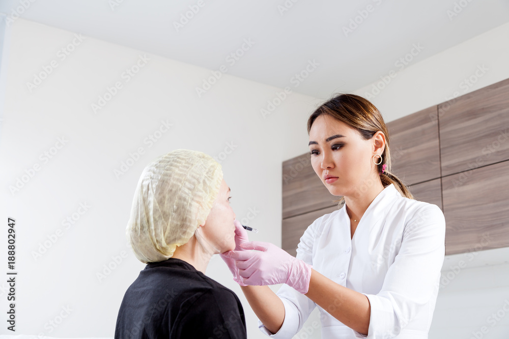 Obraz premium Young woman doctor beautician in white lab coat and sterile gloves draws a marking on woman's cheekbones before injecting Botox to correct forms