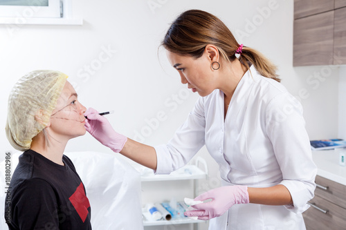 Young woman doctor beautician in white lab coat and sterile gloves draws a marking on woman s cheekbones before injecting Botox to correct forms