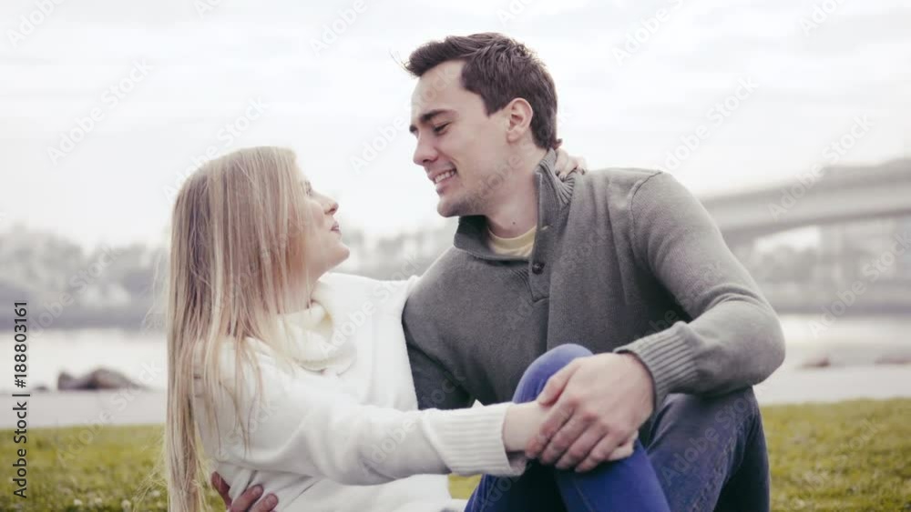 Couple in love sitting on the grass and hugging in a park. Against the background of a beautiful bay and a bridge. Pleasant evening near the ocean. Beautiful smiles. Talking and kissing. Couple in love sitting on the grass and hugging in a park. Against the background of a beautiful bay and a bridge. Pleasant evening near the ocean. Beautiful smiles. Talking and kissing.
